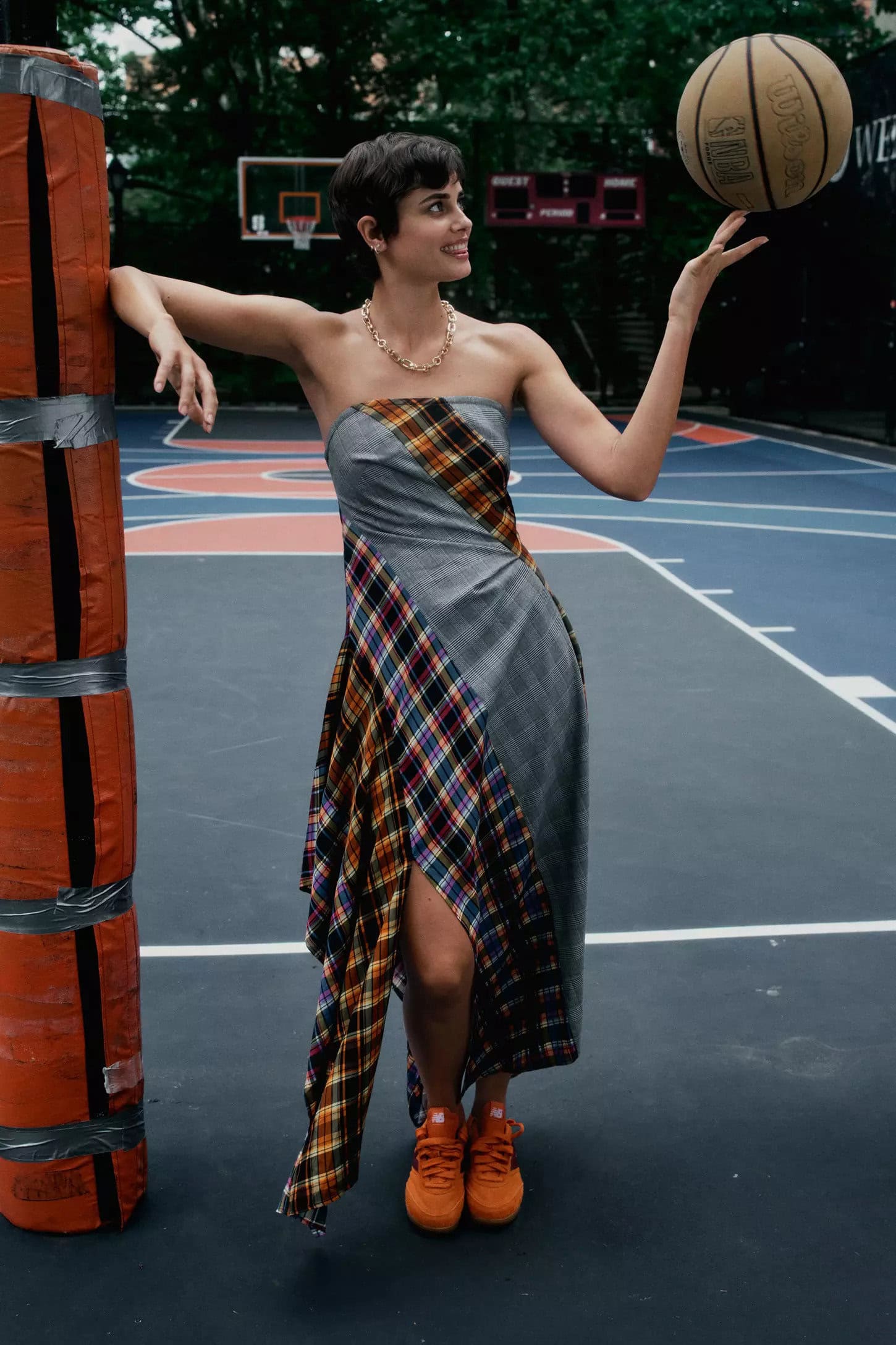 Woman with short dark hair in a plaid dress balancing a basketball on her finger on a blue outdoor basketball court