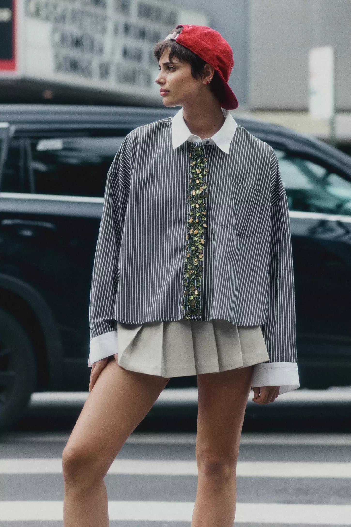Model wearing a striped button-down shirt, pleated skirt, and red cap standing outdoors in New York City.