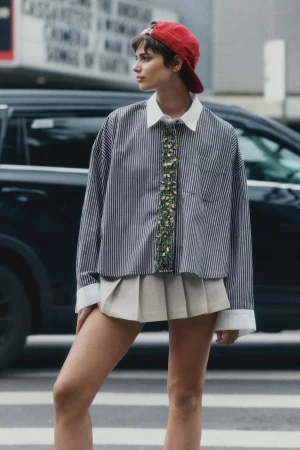 Model wearing a striped button-down shirt, pleated skirt, and red cap standing outdoors in New York City.