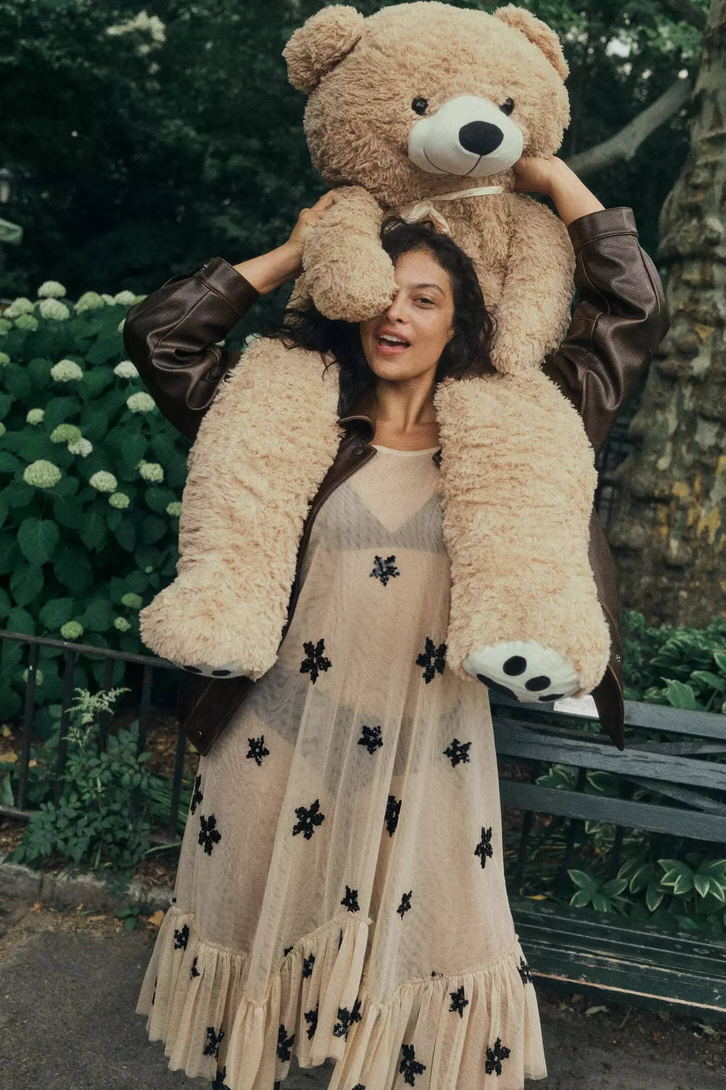 Model carrying a giant teddy bear on her shoulders, wearing a sheer floral dress and brown leather jacket in a park.