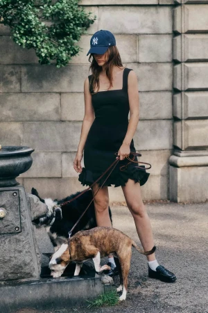 Woman wearing a blue baseball cap and black dress standing outdoors with two dogs drinking from a stone fountain