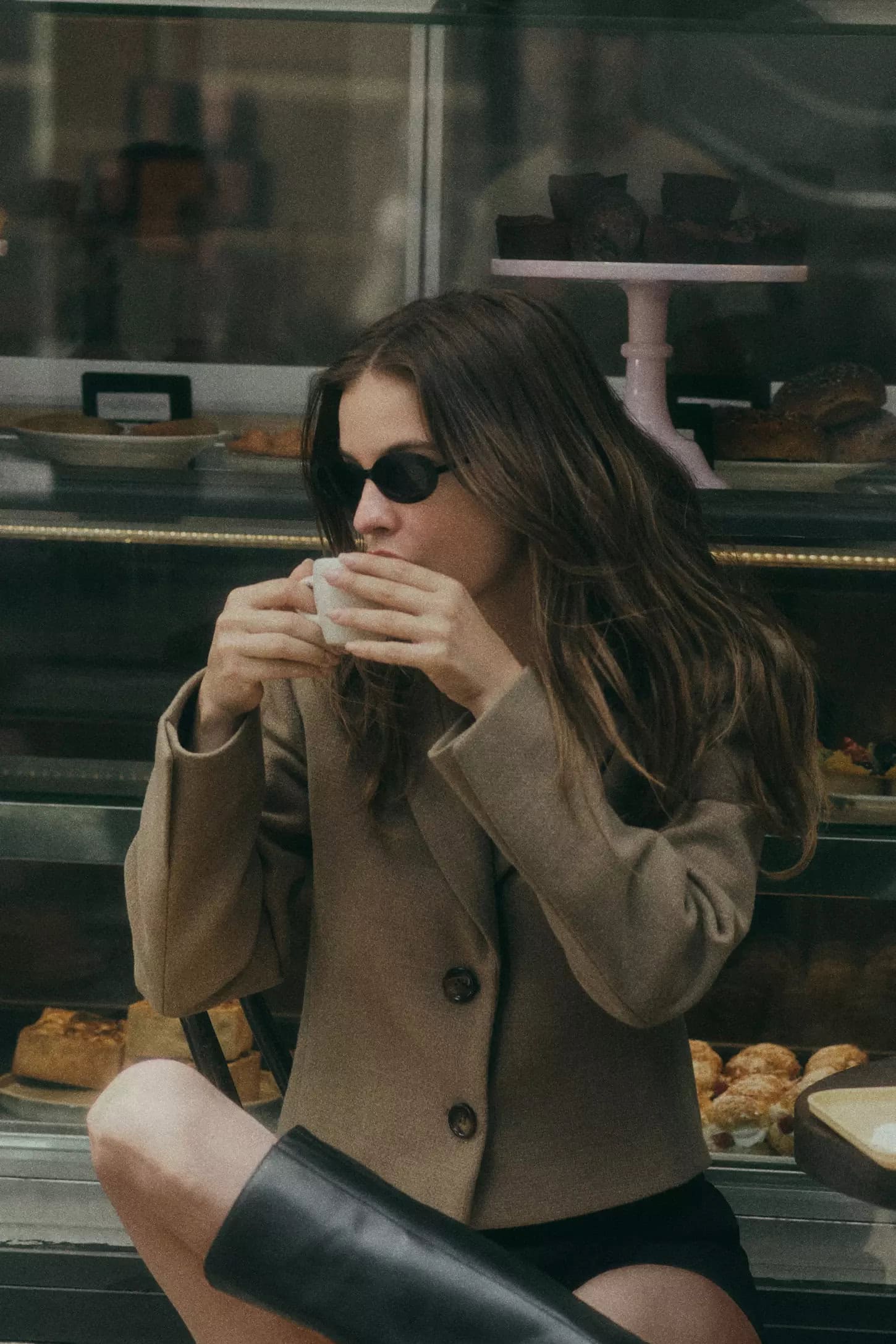 Model wearing dark sunglasses and a camel jacket drinking from a small white cup in front of a café display case