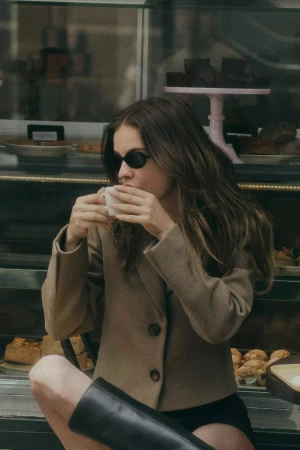 Model wearing dark sunglasses and a camel jacket drinking from a small white cup in front of a café display case
