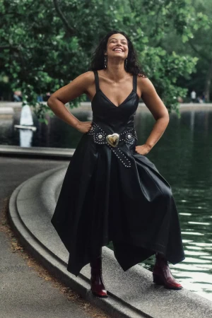 Woman in a black leather dress with a heart buckle belt and red boots posing outdoors by a park pond