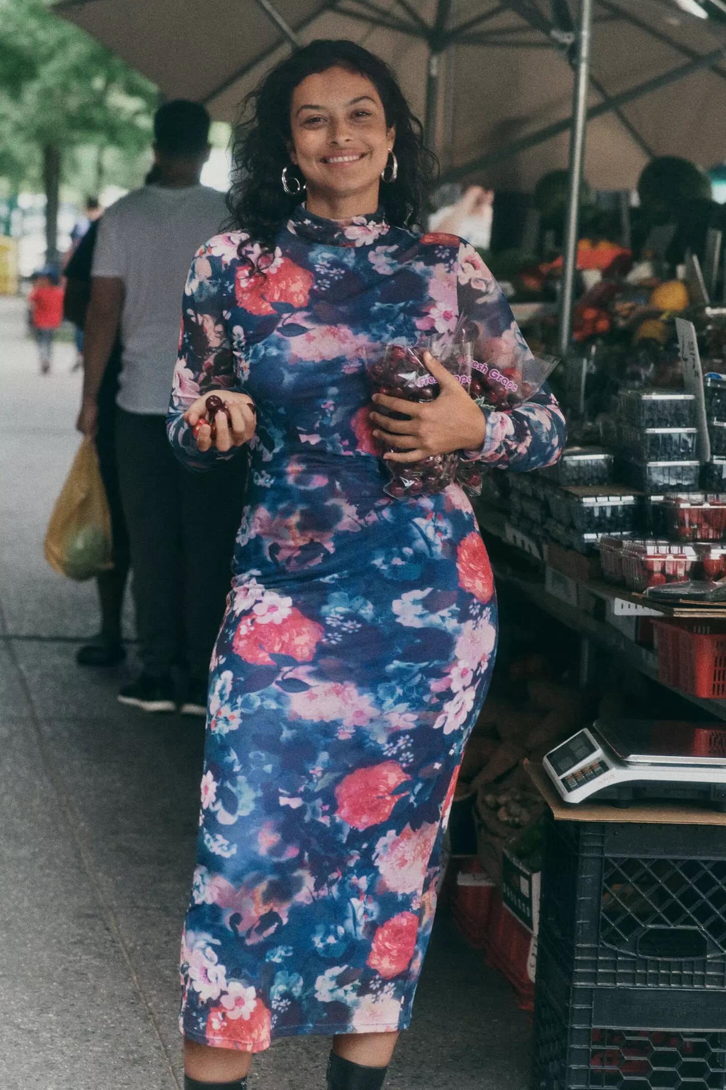 Smiling woman wearing a floral long-sleeve midi dress holding produce at an outdoor market stall.