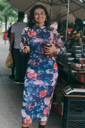 Smiling woman wearing a floral long-sleeve midi dress holding produce at an outdoor market stall.