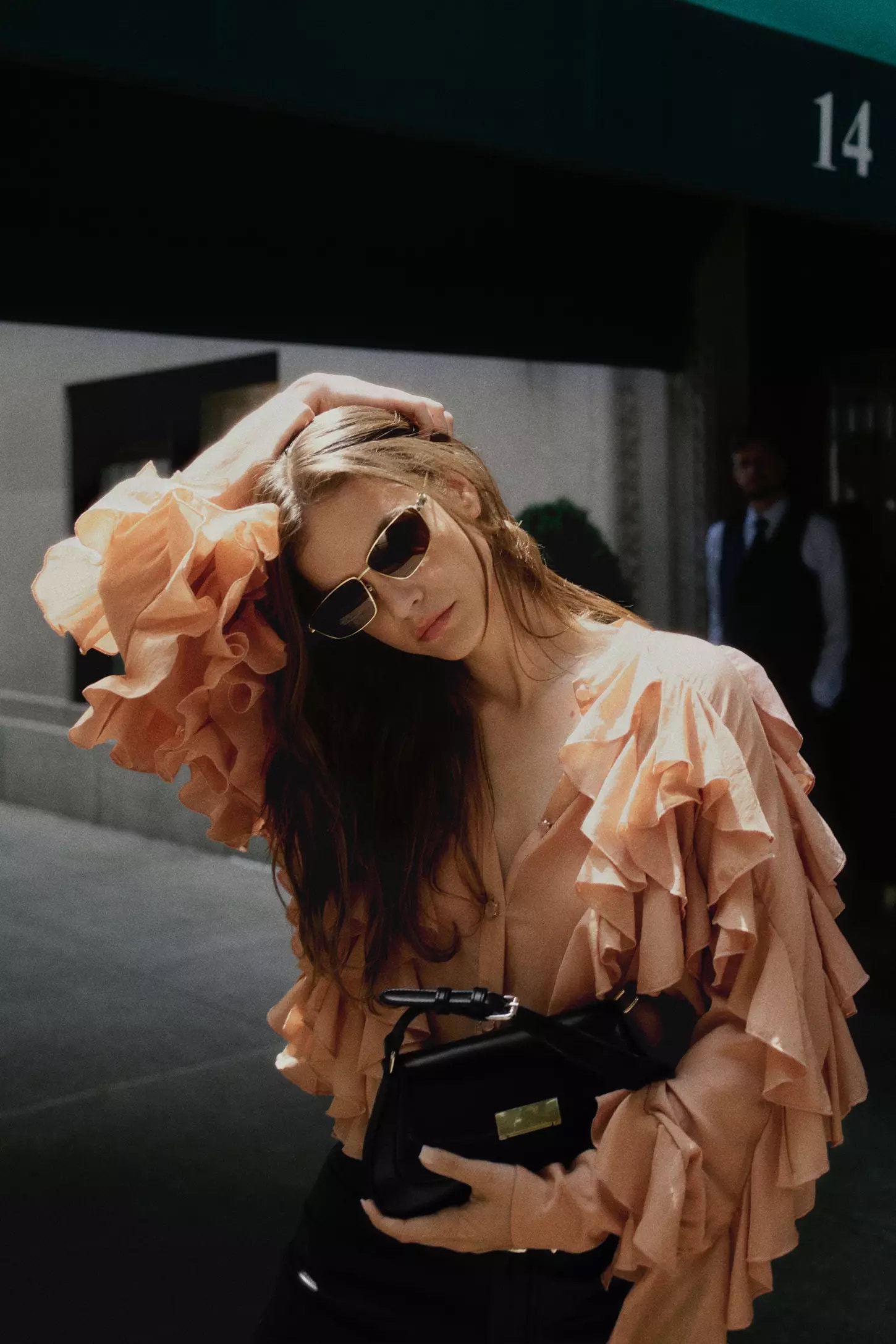 Model in peach ruffled blouse holding a black handbag and wearing sunglasses against an urban storefront.