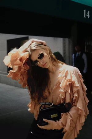 Model in peach ruffled blouse holding a black handbag and wearing sunglasses against an urban storefront.