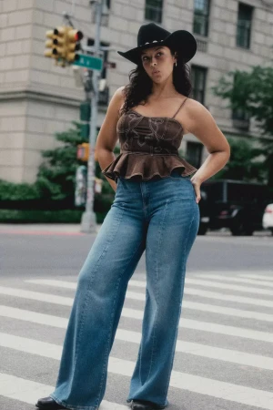 Model wearing a brown peplum top, blue jeans, and a black cowboy hat standing on a city street.