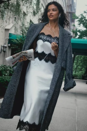 Woman walking outdoors wearing a white satin slip dress with black lace trim and a herringbone coat, holding a newspaper.