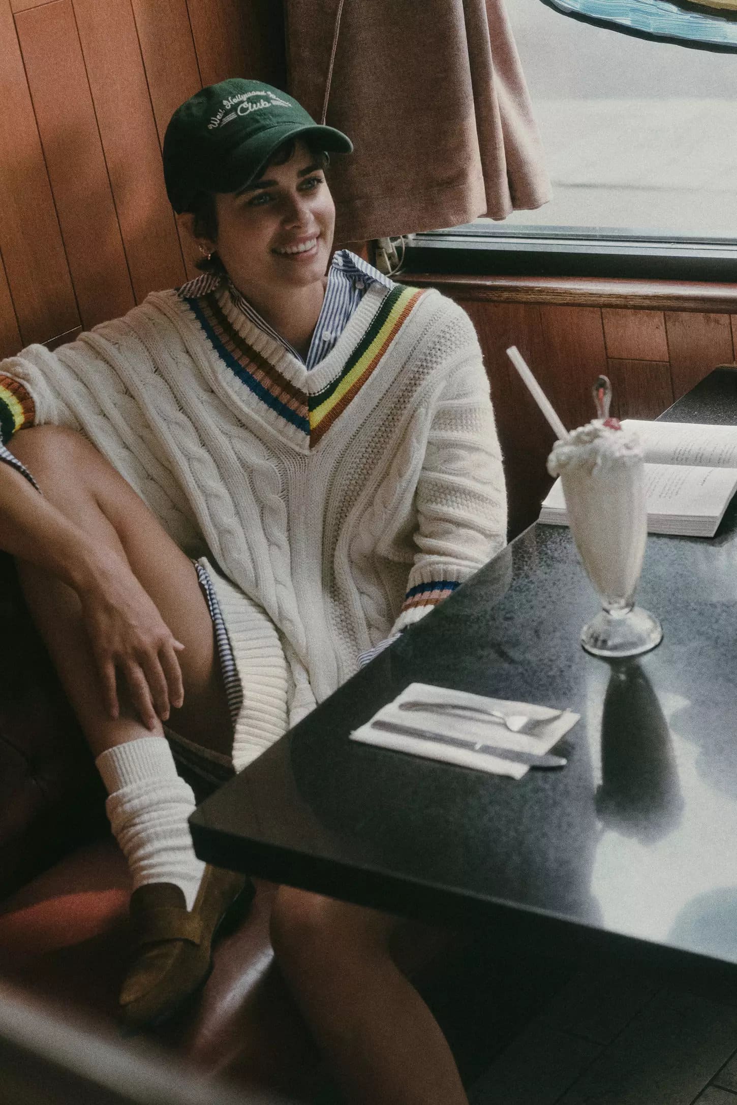 Woman wearing a green baseball cap and a cream cable-knit sweater seated at a diner booth next to a milkshake