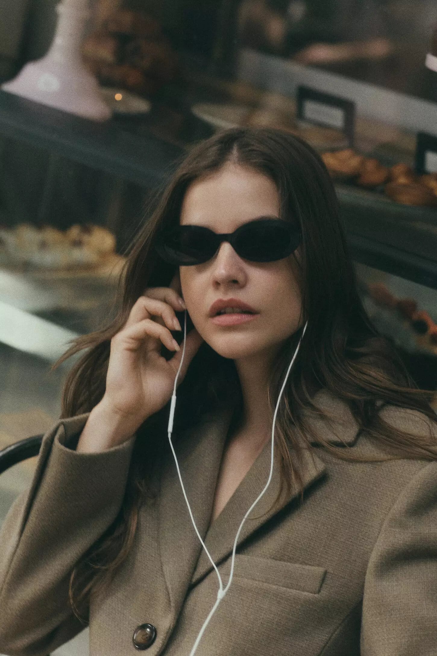 Model Barbara Palvin wearing black sunglasses and a brown coat, holding wired earbuds inside a cafe for Anthropologie.