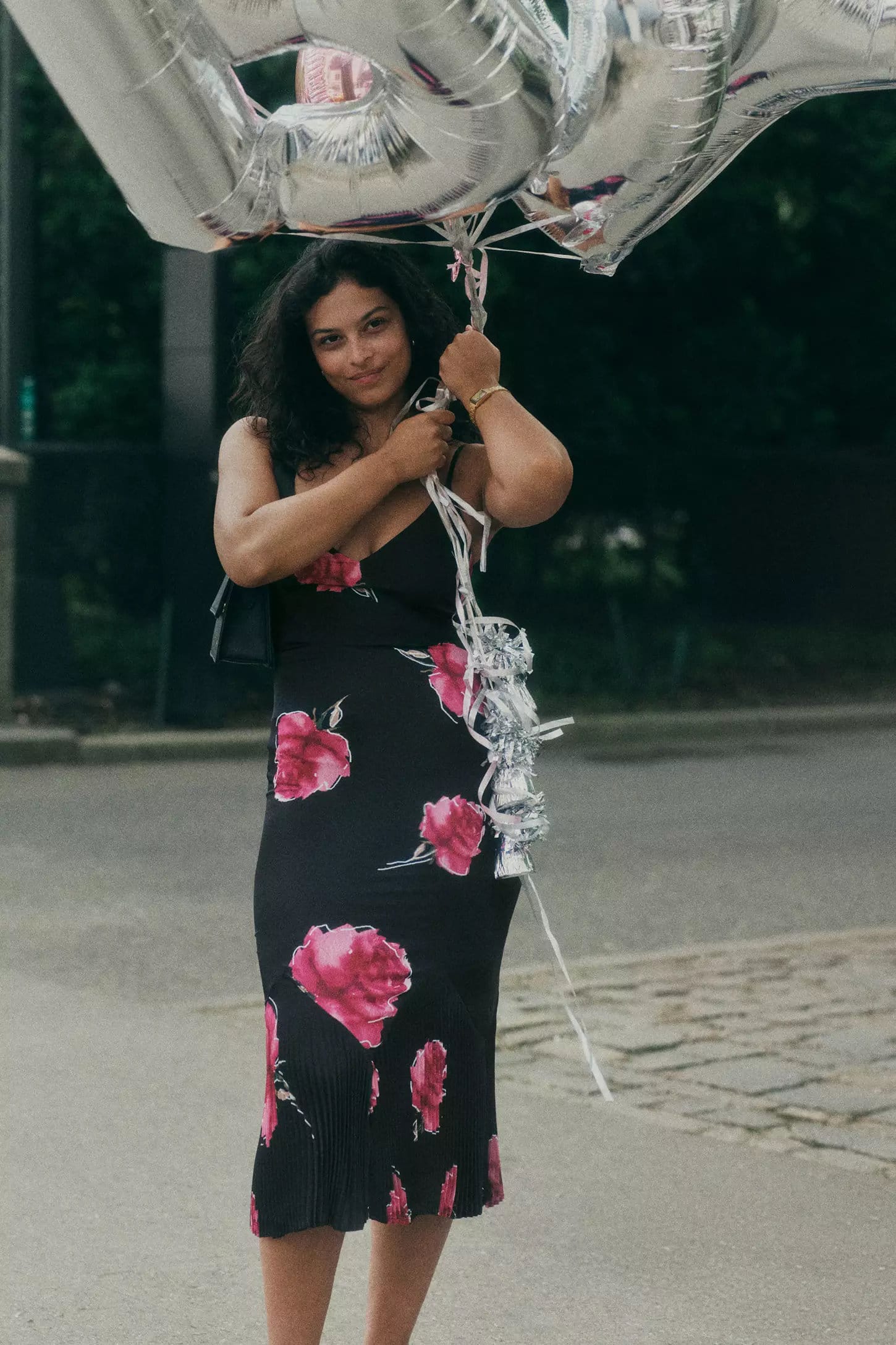 Woman in a black floral dress holding large silver number balloons on an outdoor street