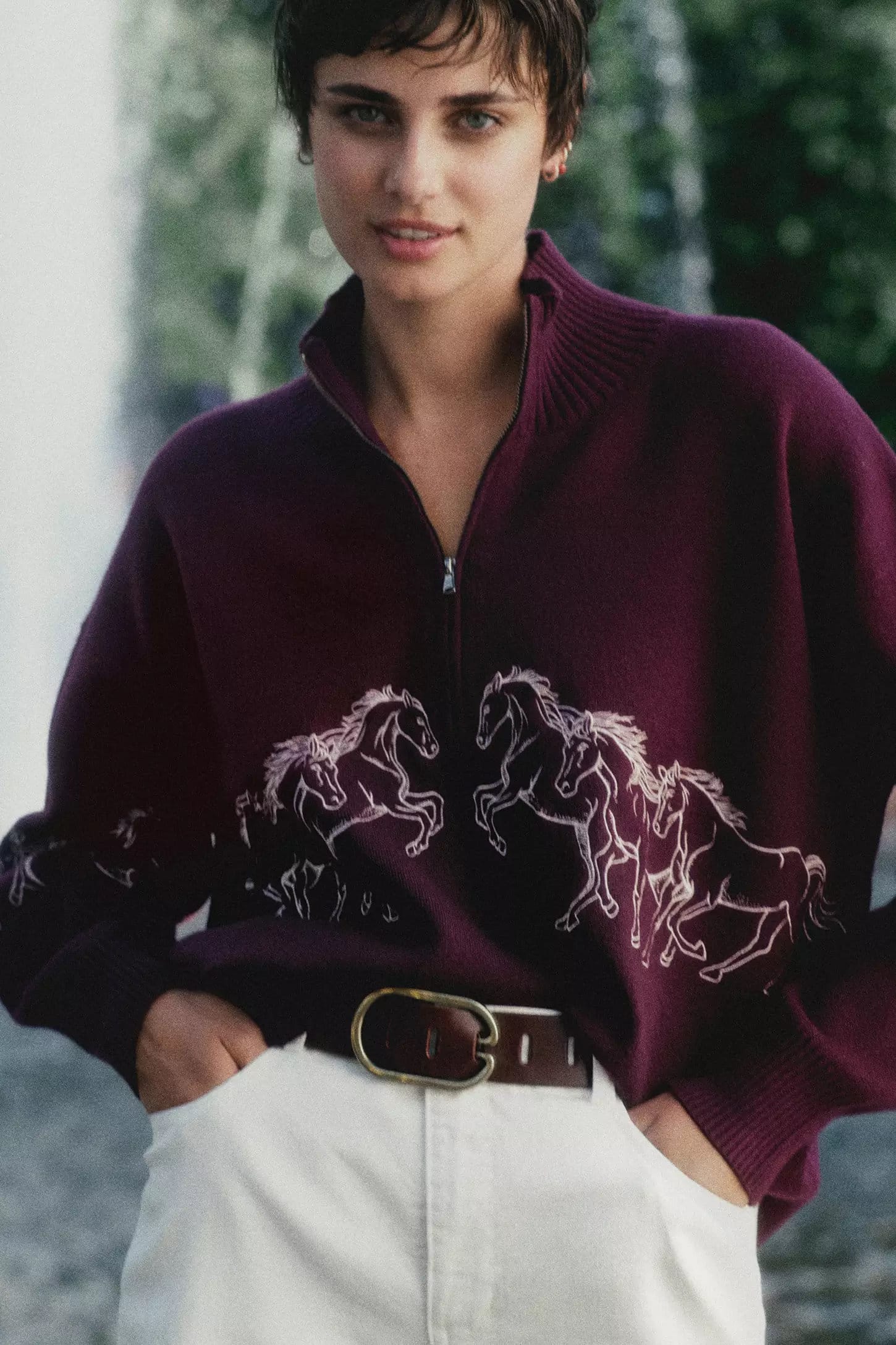 A woman with short dark hair wearing a burgundy zip-up sweater with embroidered white horses and white pants.