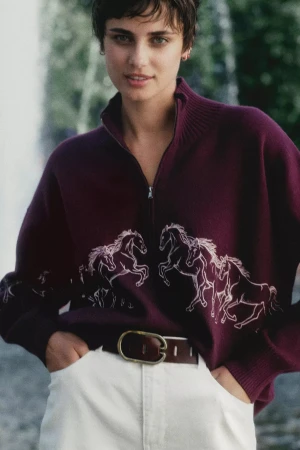 A woman with short dark hair wearing a burgundy zip-up sweater with embroidered white horses and white pants.