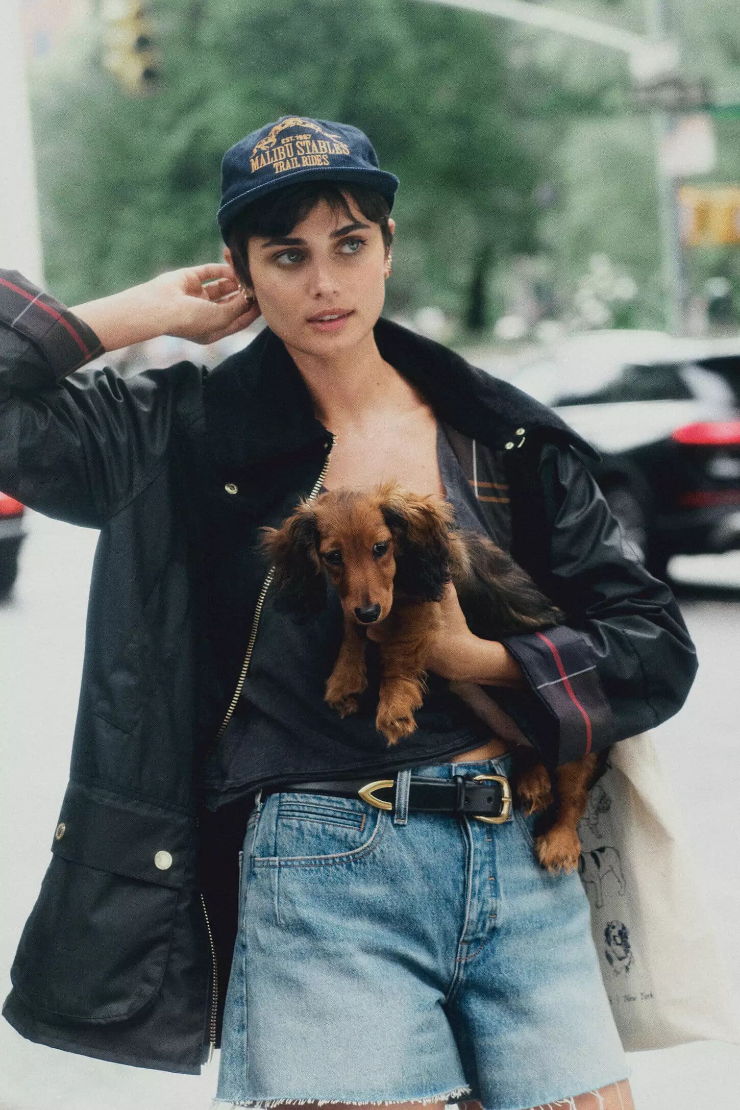 Model Taylor Hill wearing a dark baseball cap and jacket holding a long-haired dachshund dog on a New York City street