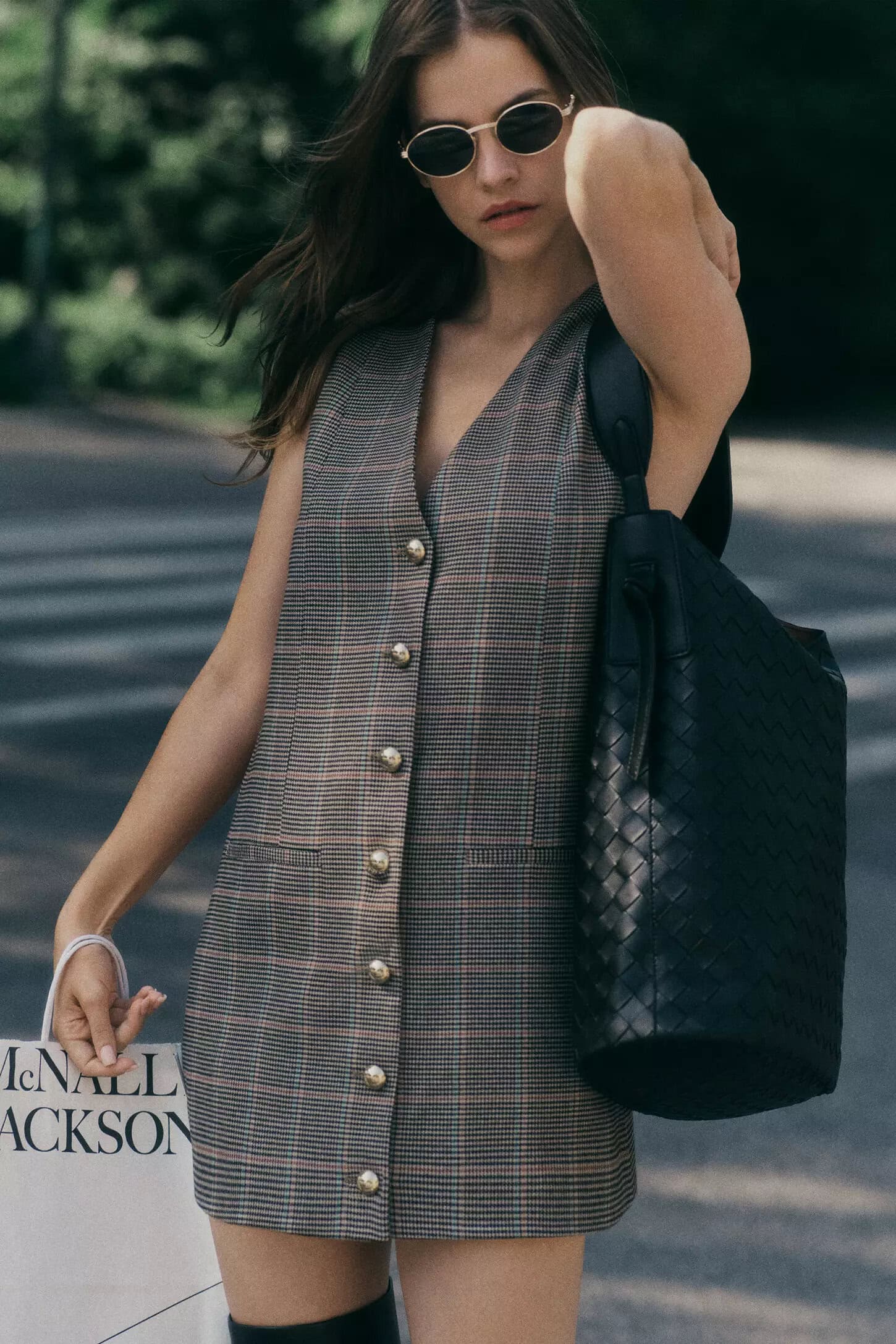 Barbara Palvin wearing a plaid button-front vest and dark sunglasses holding a black leather tote bag outdoors