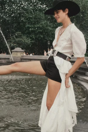 Model in white blouse and black shorts wearing a cowboy hat near a park fountain