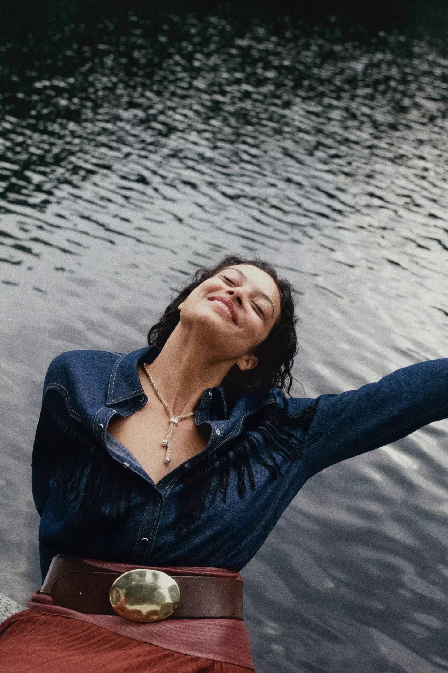 Smiling woman in a denim shirt with fringe and a wide brown belt posing against dark rippling water
