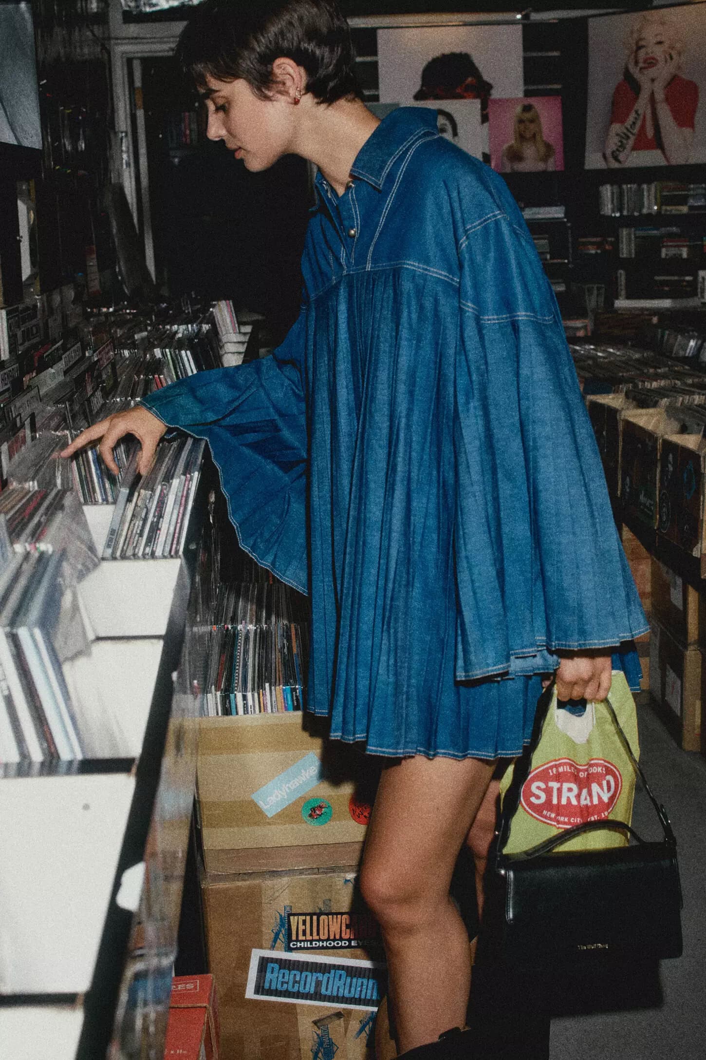 Short-haired woman browsing records in a music store wearing an oversized blue denim dress and holding a black handbag.