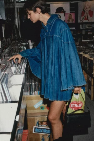 Short-haired woman browsing records in a music store wearing an oversized blue denim dress and holding a black handbag.