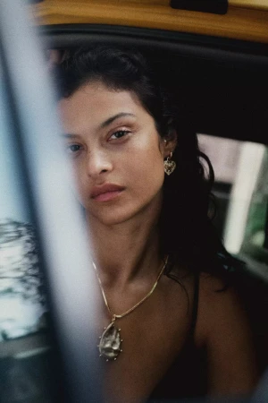 Close-up portrait of a woman wearing gold pendant necklace and earrings looking out from inside a car