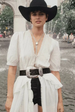 Model wearing a black cowboy hat, white blouse, and statement belt standing outdoors near a fountain.