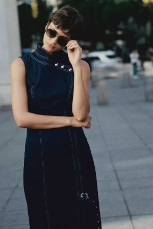 Model with short hair wearing sunglasses and a dark denim dress standing outdoors on a paved street