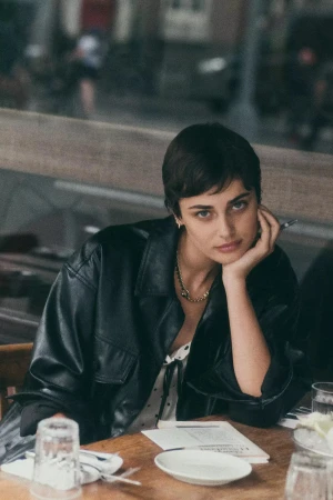 Taylor Hill with short dark hair in a black leather jacket resting chin on hand at a wooden cafe table