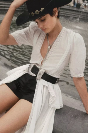 Taylor Hill wearing a white lace blouse, black shorts, and cowboy hat with silver buckle belt outdoors for Anthropologie.