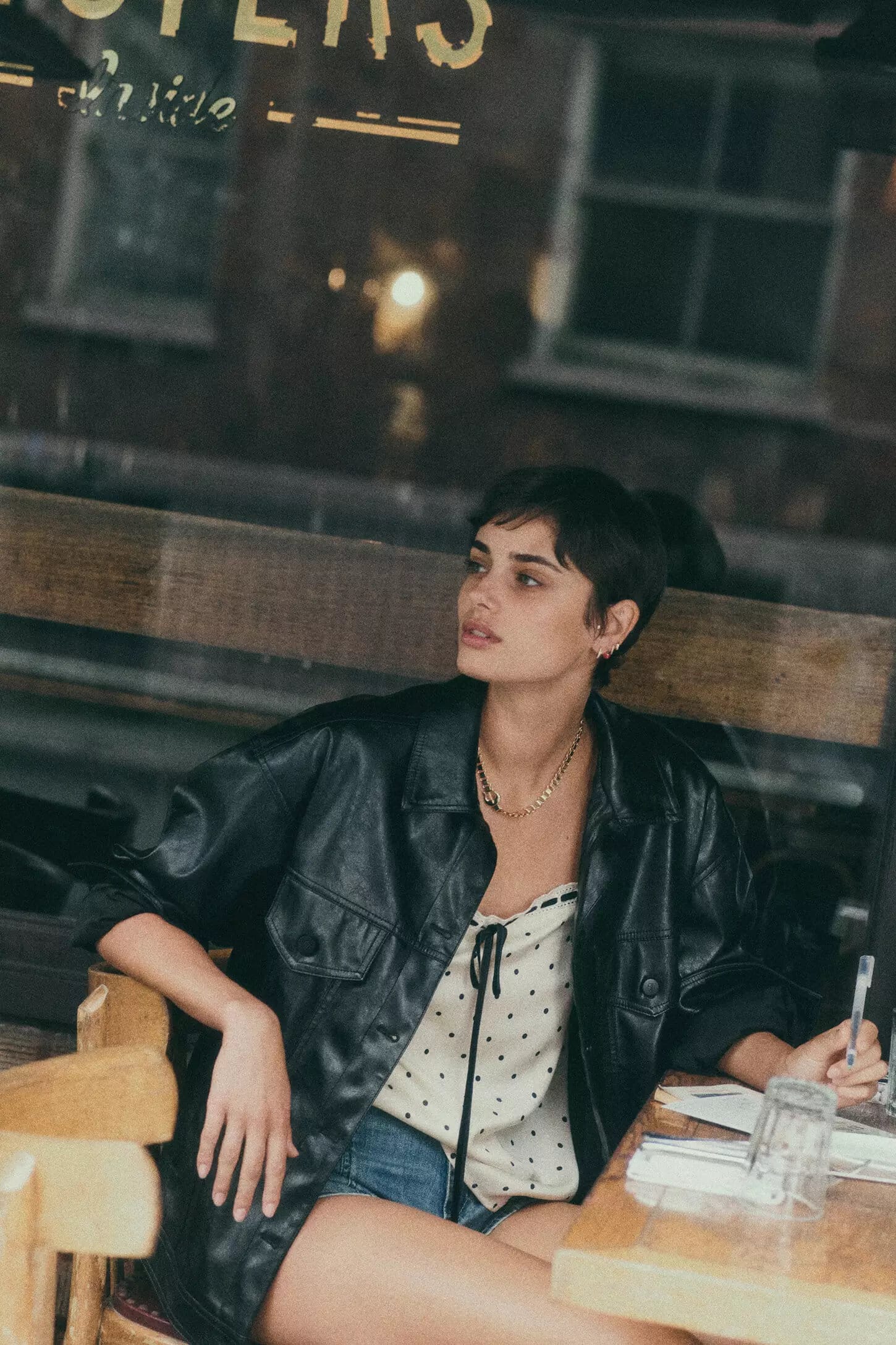 Model Taylor Hill seated at a cafe table wearing a black leather jacket and patterned top