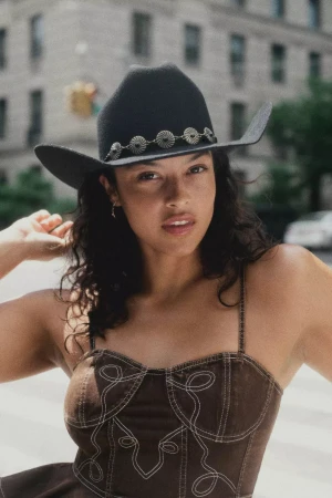 Model wearing a black cowboy hat with silver concho band and a brown top outdoors in New York City for Anthropologie.