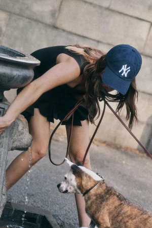 Woman in a dark blue baseball cap leaning over a stone fountain to let her brindle dog drink water.