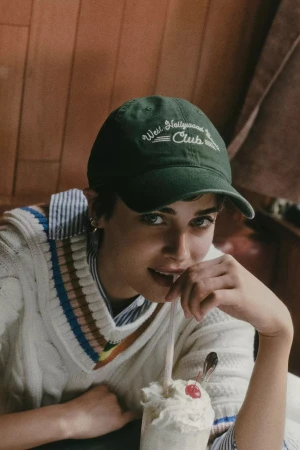 Woman wearing a green baseball cap and cream cable-knit sweater sipping a milkshake with a cherry on top.
