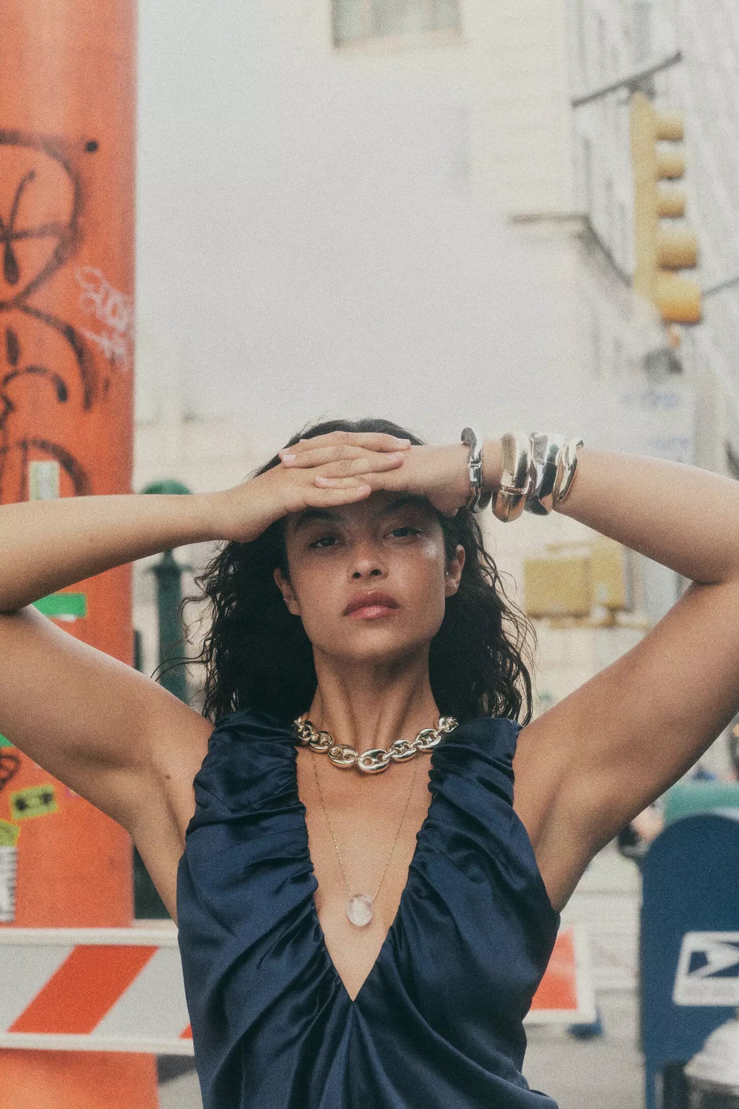 Woman in blue dress wearing a chunky gold chain necklace holding hands to her forehead on a city street.