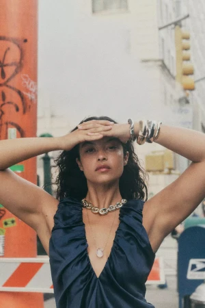 Woman in blue dress wearing a chunky gold chain necklace holding hands to her forehead on a city street.