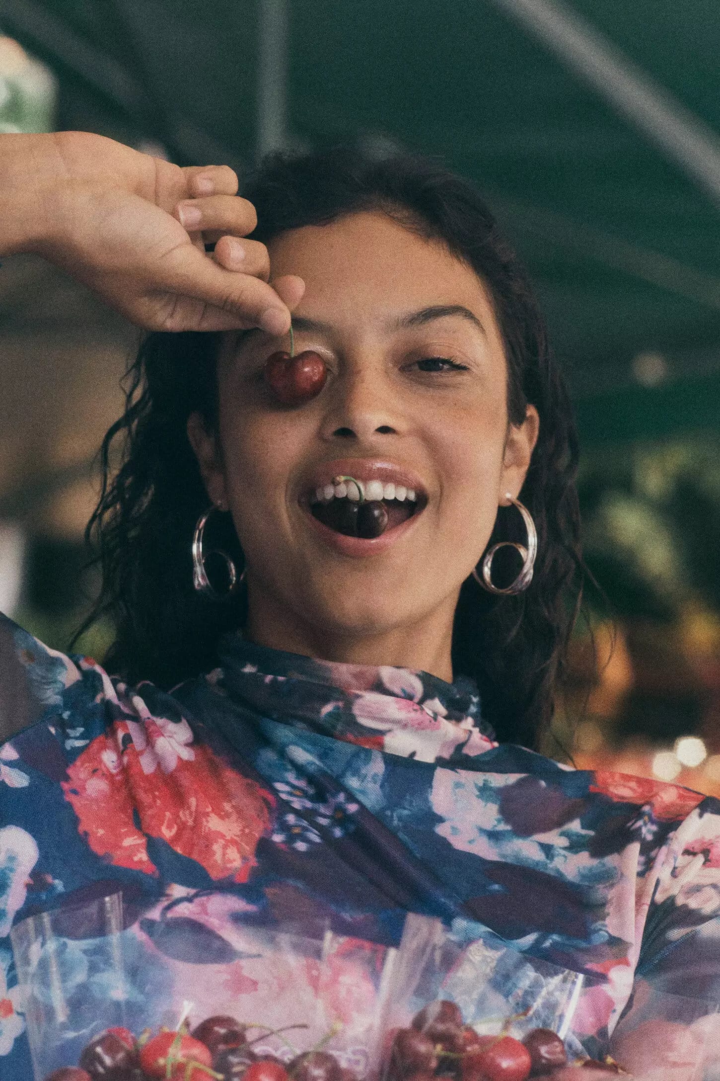 Woman holding cherries in front of her eye and smiling while wearing a floral top and silver hoop earrings