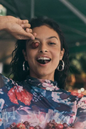 Woman holding cherries in front of her eye and smiling while wearing a floral top and silver hoop earrings