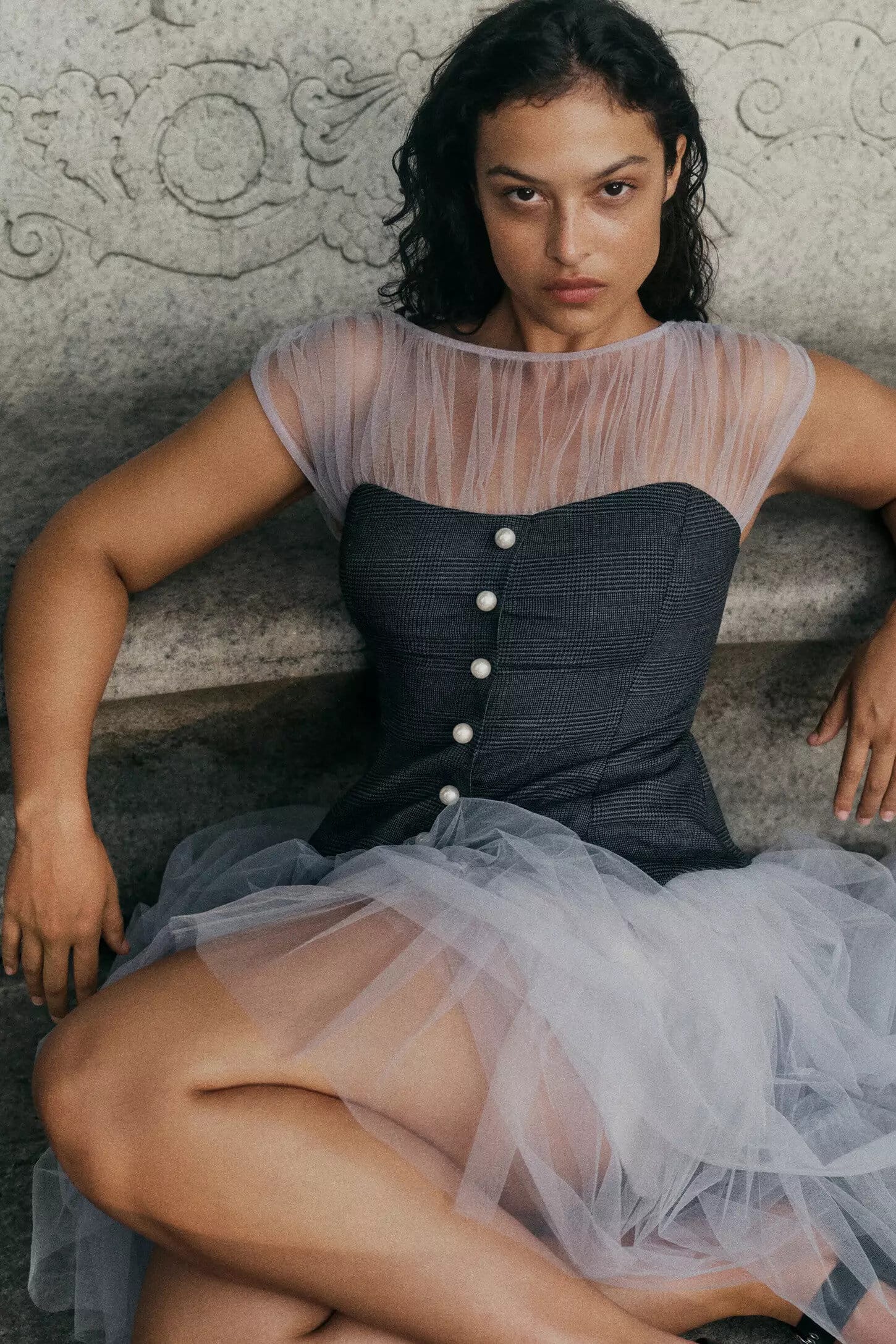 Model wearing a grey structured corset top with tulle accents seated against a carved stone architectural backdrop.