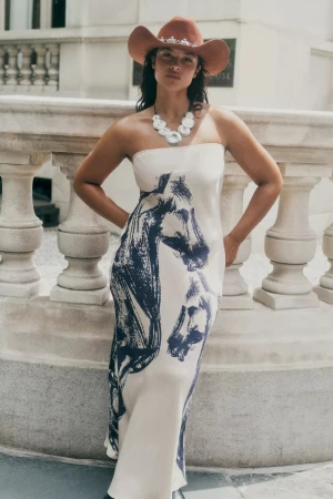 Model wearing a strapless printed dress and brown cowboy hat posing against an ornate stone balustrade