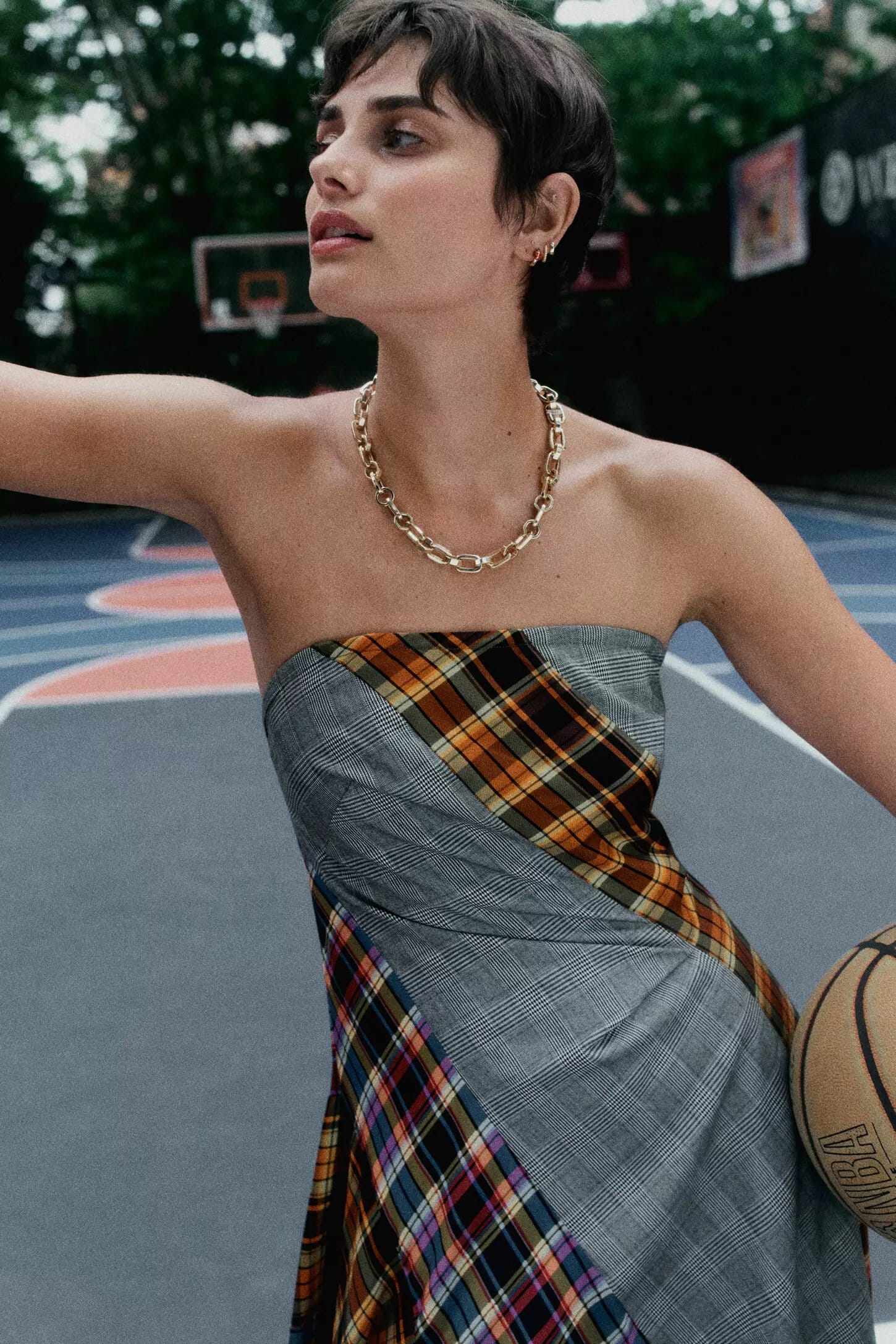 Model with short dark hair wearing a plaid strapless dress and gold chain necklace holding a basketball on a blue outdoor court