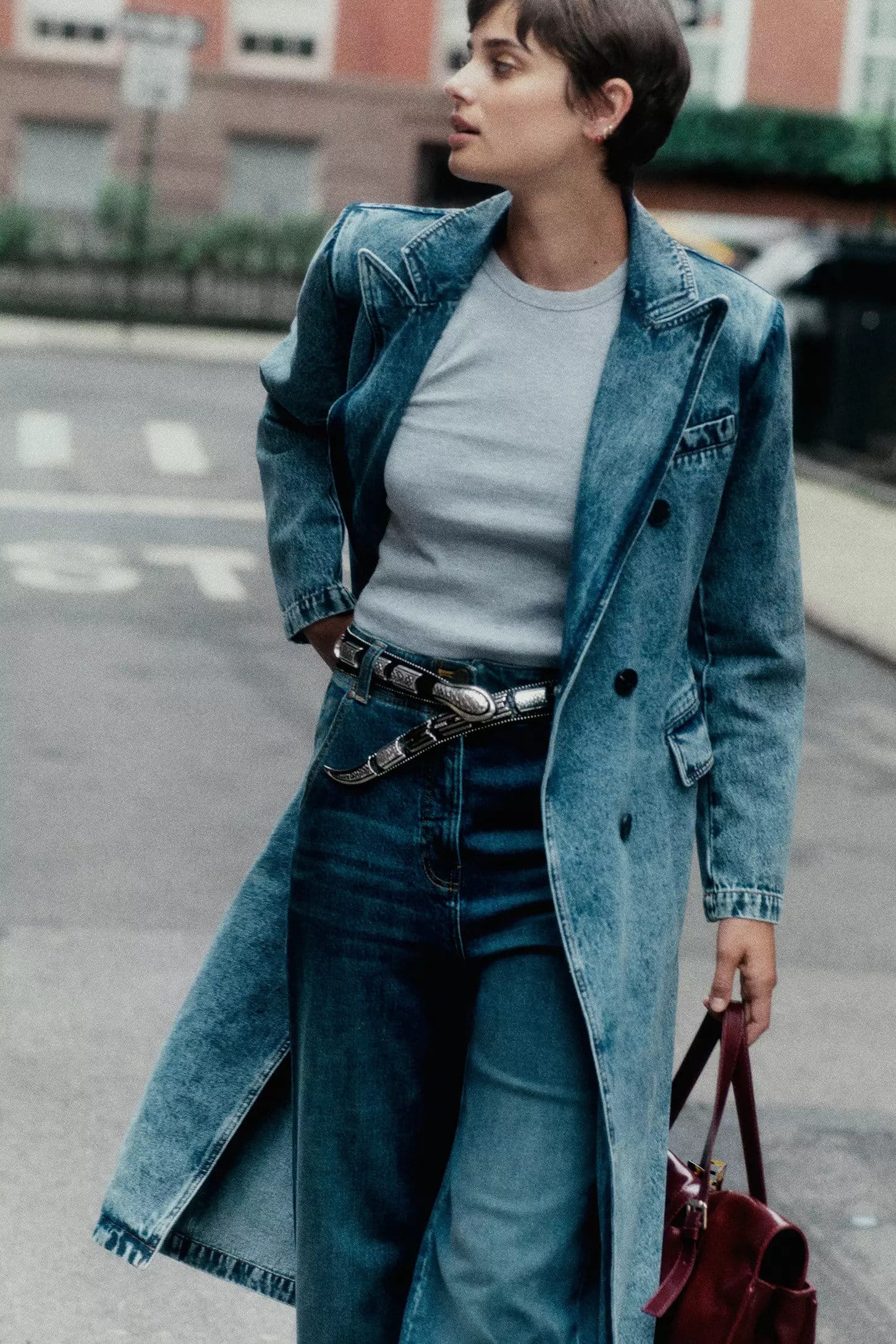 Taylor Hill in a long denim coat, jeans, and belt walking on a city street holding a handbag