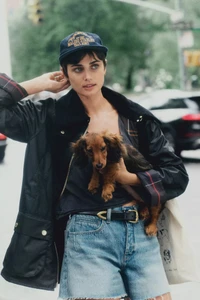 Model Taylor Hill wearing a dark baseball cap and jacket holding a long-haired dachshund dog on a New York City street