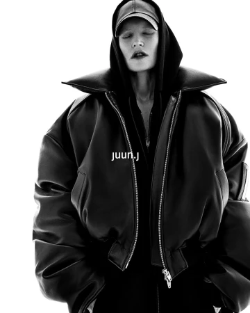Model wearing an oversized black leather bomber jacket and a baseball cap against a bright white background.