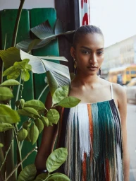 Model in a multicolored fringed dress posing beside lush green foliage with a city street background