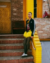 Model standing on brick building steps wearing a black track jacket and yellow feathered skirt