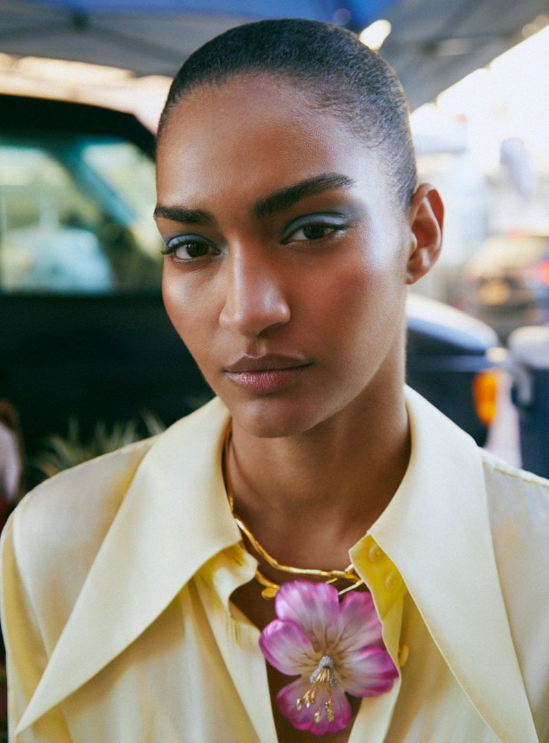 Portrait of a woman with slicked-back hair and a large flower necklace, wearing a pale yellow blouse in an outdoor setting.