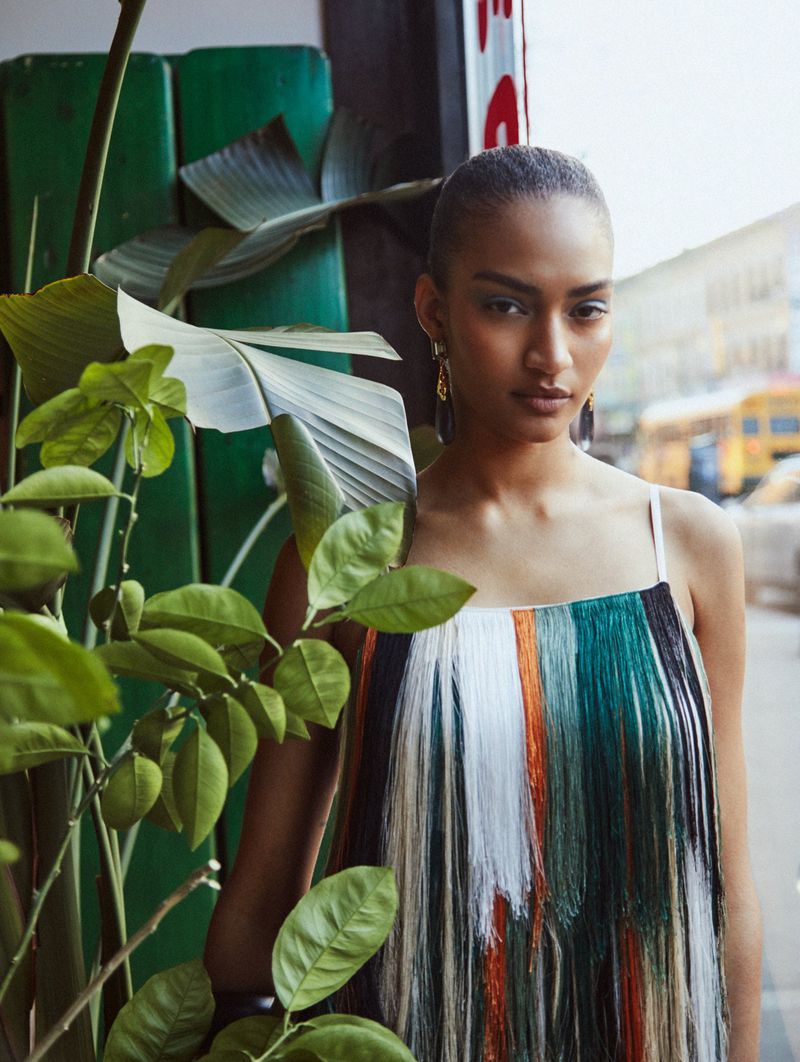Portrait of a woman in a fringed dress standing by a green wall with plants, featuring an urban street scene in the background.