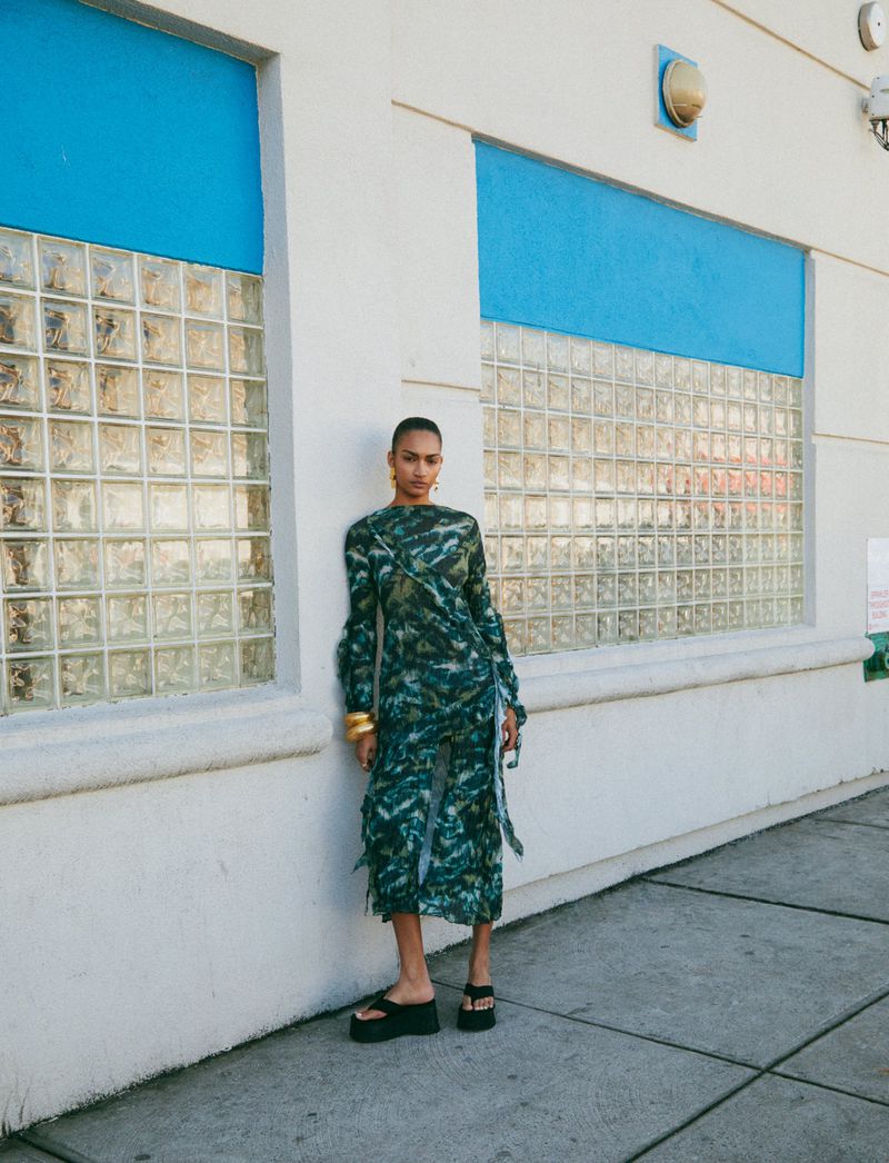 A woman poses in a green patterned dress and platform sandals in an urban setting, standing against a white wall with blue trim.