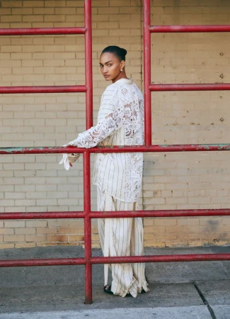 Woman in a white lace-back top and striped pants, posing by a red metal barrier with a brick wall background in an outdoor setting.